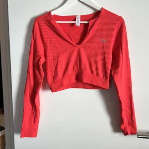 Röd ribbad tröja från ICANIWILL - Säljer en snygg korallröd ribbad tröja från ICANIWILL. Den har en v-ringad design och är croppad, vilket gör den perfekt för både träning och vardag. Tröjan är långärmad och tillverkad i ett stretchigt material för bästa passform. Perfekt för dig som vill ha en stilren och bekväm look!