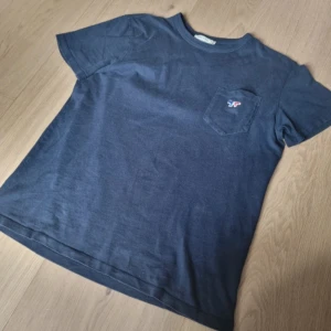 Maison Kitsuné T-shirt Mörkblå - Säljer en snygg och nästan oanvänd Maison Kitsuné t-shirt i storlek S. Den är mörkblå med den klassiska loggan på bröstfickan. Nypris var 1500 kr, men jag säljer den för 350kr. Perfekt för dig som vill ha kvalitet och stil i garderoben