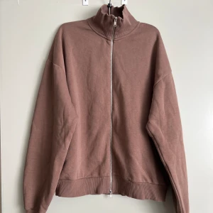 Brun zip-up tröja - Säljer en mysig brun zip-up tröja med hög krage. Den har långa ärmar och ribbade muddar vid ärmslut och nederkant. Perfekt för en avslappnad stil!