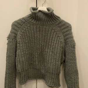Grå stickad polotröja - Säljer en mysig grön stickad polotröja med lång ärm. Tröjan har en hög krage och är perfekt för kyliga dagar. Den har en liten skada på ena ärmen. Perfekt för höst och vinter!