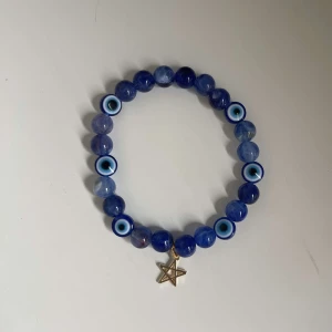 Blått armband med ögonmotiv och stjärnhänge - Säljer ett coolt blått armband med runda pärlor och ögonmotiv. Armbandet har ett litet guldstjärnhänge som ger det en unik touch. Perfekt för att lägga till lite mystik till din stil!