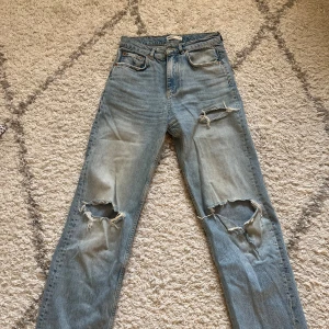 Blå slitna jeans - Säljer ett par blå jeans med slitningar på knäna. De har en klassisk femficksdesign och en ljus tvätt som ger en vintagekänsla. Perfekta för en avslappnad stil!