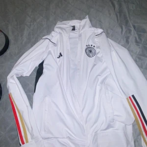 Svarta träningsbyxor med Tysklands landslagsmärke - Säljer ett par svarta träningsbyxor och tröjan  med Tysklands landslagsmärke. Byxorna har ränder i rött, gult och svart längs sidorna och är perfekta för fotbollsträning. De är i bra skick och har en bekväm passform.