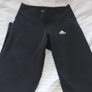 Svarta leggings från Adidas - Säljer ett par svarta leggings från Adidas i storlek dam S. De är högmidjade och har en tight passform, perfekta för träning. Materialet är syntetiskt och har ett ribbat mönster på sidorna. Det finns en liten reva på ena benet, men annars i bra skick.