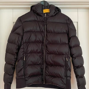 Svart Parajumpers Pharrell jacka  - Svart dunjacka med huva. 100 % dun. Yttermaterialet, polyester-nylon, är vattenavvisande och har god andningsförmåga. Framtill finns två fickor med dragkedjor. Inuti jackan finns en ficka. Storlek Y-M = 14 år. Längd 59 cm, skuldermått 43 cm. 
