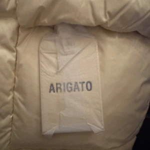 Beige dunväst från Axel Arigato - Säljer en snygg beige dunväst från Axel Arigato. Västen är perfekt för höst och vinter med sin varma och puffiga design. Den har en stilren look med knappar framtill och en diskret logga. Passar perfekt för både vardag och festliga tillfällen.