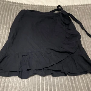 Svart omlottkjol med volang - Snygg svart omlottkjol med volangdetaljer. Kjolen har en knytfunktion i midjan för justerbar passform. 