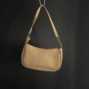 Beige axelväska med krokodilmönster - Snygg beige axelväska med krokodilmönster och guldfärgade kedjedetaljer. Väskan har en dragkedja upptill och ett rymligt innerfack. Perfekt för att ge din outfit en trendig touch.