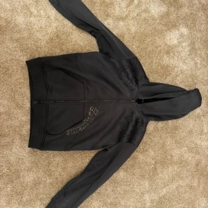 Svart hoodie från Chili - Säljer en svart hoodie från Chili i storlek 130. Den har en dragkedja framtill och är tillverkad i 100% polyester. Perfekt för en avslappnad stil med snyggt mönster på bröstet.