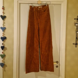 Orangebruna manchesterbyxor från Levi's - Säljer ett par snygga orangebruna manchesterbyxor från Levi's med hög midja och breda ben. Byxorna har klassiska femficksdetaljer och en knappgylf. Perfekta för en trendig och avslappnad stil.