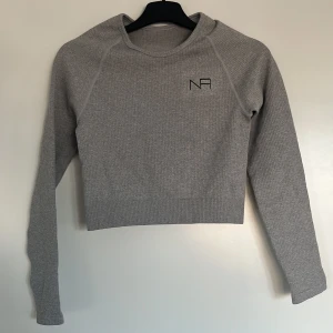 Grå ribbad croppad tröja från NA - Säljer en stilren grå ribbad tröja från NA. Den är croppad och har långa ärmar, perfekt för en trendig look. Tröjan har en diskret logga på bröstet. Passar bra till både jeans och kjol.