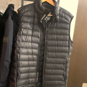 Svart dunväst från 70 Degrees North TENSON - Säljer en svart dunväst från 70 Degrees North. Västen har en dragkedja framtill och är quiltad för extra värme. Perfekt för kyligare dagar och ger en stilren look.