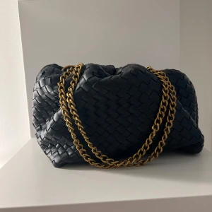 Svart flätad axelväska med guldkedja - Snygg svart axelväska i flätat skinn med en elegant guldkedja som axelrem. Väskan har en stilren design och är perfekt för att ge en lyxig touch till din outfit. Den rymliga insidan gör den praktisk för vardagsbruk.