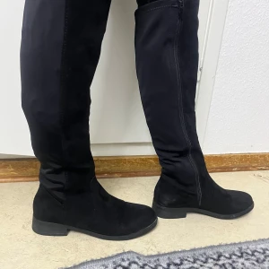 Svarta höga stövlar med dragkedja - Snygga svarta stövlar i mocka med dragkedja på insidan. De är knähöga och har en låg klack, vilket gör dem både stiliga och bekväma. Perfekta för höst- och vinterdagar.