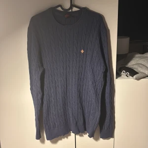 Blå kabelstickad tröja från Morris - Säljer en stilren blå kabelstickad tröja från Morris. Tröjan har en klassisk rund halsringning och ett diskret emblem på bröstet. Perfekt för kyligare dagar och ger en tidlös look.