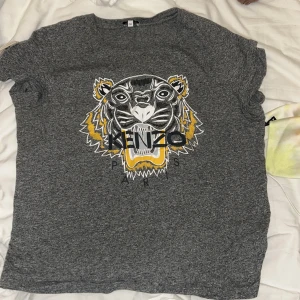 Grå t-shirt med tigertryck från Kenzo - Snygg grå t-shirt från Kenzo med ett stort tigertryck i svart, vitt och gult på framsidan. Perfekt för en avslappnad stil med en touch av attityd. T-shirten har korta ärmar och en rund halsringning. Äkta vara! 