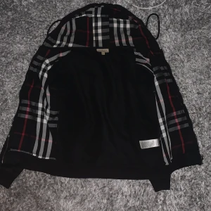 Svart kofta från Burberry - Säljer en stilren svart kofta från Burberry med klassiskt rutigt foder. Koftan har en dragkedja framtill och en broderad logotyp på bröstet. Perfekt för dig som vill ha en tidlös och elegant look.