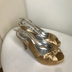 Guldiga klackskor från Luana - Eleganta guldiga klackskor från Luana med öppen tå och dekorativ rosett framtill. Skorna har en smal rem runt hälen och en stilren klack som ger en sofistikerad look. Perfekta för speciella tillfällen. Som student eller annat 