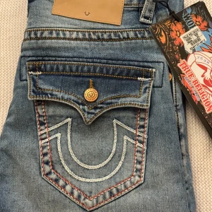 Blå jeans från True Religion - Snygga blå jeans från True Religion med slitna detaljer och kontrastsömmar. De har en unik bakficka med broderad logga och knappstängning. Perfekta för en avslappnad stil.