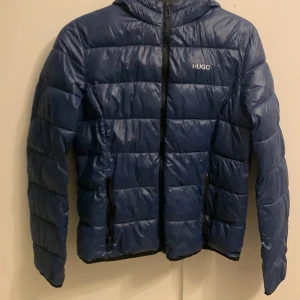 Blå dunjacka från Hugo Boss - Säljer en snygg blå dunjacka från Hugo Boss. Jackan har en quiltad design med dragkedja framtill och en praktisk huva. Perfekt för kyligare dagar och ger en stilren look. Passar både till vardags och mer uppklädda tillfällen.