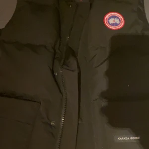 Svart dunväst från Canada Goose - Säljer en svart dunväst från Canada Goose med det klassiska Arctic Program-emblemet på bröstet. Västen har en dragkedja framtill och är perfekt för kyliga dagar. Den är tillverkad i Kanada med högkvalitativa material. Passar m