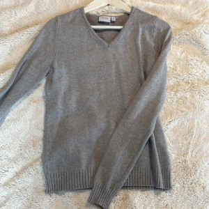 Grå V-ringad tröja  - Säljer en stilren grå V-ringad tröja från Vila. Tröjan har långa ärmar och ribbstickade muddar vid ärmslut och nederkant. Perfekt för kyligare dagar och enkel att matcha med olika outfits.