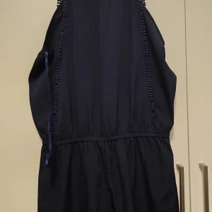 Mörkblå klänning med spetsdetaljer - Elegant mörkblå klänning med v-ringning och fina spetsdetaljer längs sidorna. Perfekt för en stilren look. Klänningen har en smickrande passform och är ärmlös.