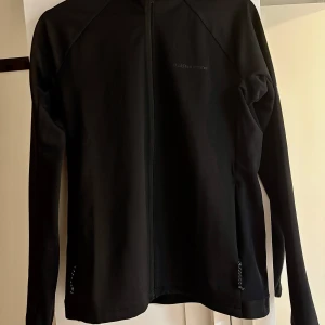 Svart jacka från Peak Performance - Snygg svart vindjacka från Peak Performance med dragkedja framtill. Jackan har en stilren design och är perfekt för en sportig look. Den är långärmad och har en diskret logga på bröstet. Helt ny och oanvänd, endast testad.