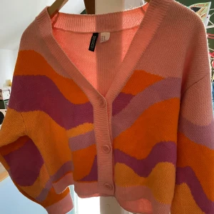 Färgglad kofta från HM - Säljer en färgglad kofta från HM med vågiga mönster i rosa, orange och lila. Koftan har en v-ringad design med knappar framtill och långa ärmar. Jag är en XS men passar också S