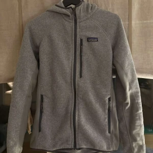 Grå fleecejacka från Patagonia - Säljer en grå fleecejacka från Patagonia med huva och dragkedja. Jackan har två sidofickor med dragkedjor och en bröstficka. Perfekt för kyliga dagar och outdoor-äventyr. Perfekt för våren och kyligare sommarkvällar! Kom privat vid funderingar!
