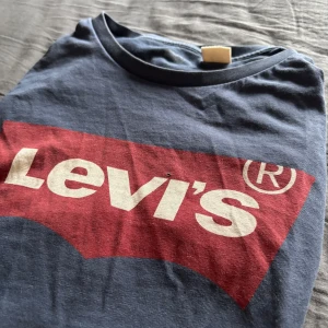 Blå t-shirt från Levi's - Säljer en blå t-shirt från Levi's med det klassiska röda och vita logotyptrycket på framsidan. T-shirten är kortärmad och tillverkad i mjukt bomullsmaterial. Perfekt för en avslappnad stil.