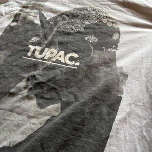 Vit t-shirt med Tupac-tryck från Mister Tee - Säljer en vit t-shirt från Mister Tee med ett stort tryck av Tupac på framsidan. T-shirten är kortärmad och har en klassisk rund halsringning. Perfekt för dig som gillar ikoniska musikmotiv.