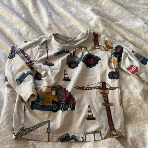 Beige långärmad tröja med byggfordon från Lindex - Säljer en beige långärmad tröja från Lindex med ett färgglatt mönster av byggfordon och trafikskyltar. Perfekt för små byggfantaster! Tröjan är i storlek 92 och har en bekväm passform. Finns en liten fläck på sista bilden men inget man tänker på