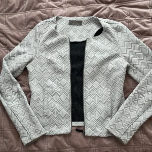 Mönstrad jacka från Vero Moda - Snygg jacka från Vero Moda med ett geometriskt mönster i vitt och grått. Jackan har en stilren design med långärmade ärmar och en dragkedja framtill. Perfekt för att ge din outfit en trendig touch.