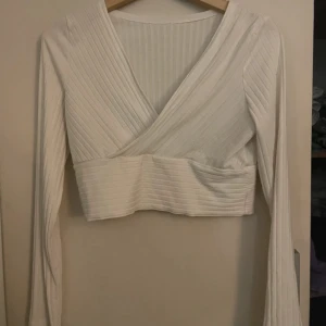 Vit ribbad omlottopp - Säljer en stilren vit ribbad omlottopp med långa ärmar. Toppen har en v-ringad design och är croppad, vilket ger en modern och trendig look. Perfekt för att matcha med höga jeans eller kjol.