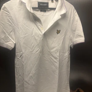 Vit pikétröja från Lyle & Scott - Säljer en stilren vit pikétröja från Lyle & Scott i slim fit. Tröjan har korta ärmar och en klassisk krage med knappar. Den är dekorerad med Lyle & Scotts ikoniska logga på bröstet. Perfekt för en avslappnad men ändå snygg look.