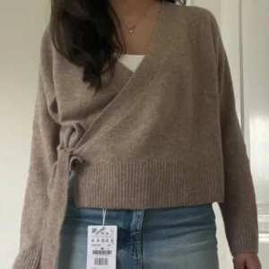 Beige omlottkofta - Säljer en mysig beige omlottkofta med knytning i sidan. Koftan har långa ärmar och ribbade kanter, perfekt för en stilren och bekväm look. Passar bra till jeans eller kjol.
