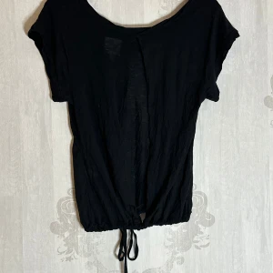 Svart kortärmad topp med knyt - Säljer en svart kortärmad topp med knyt i nederkanten. Toppen har en lös passform och är perfekt för en avslappnad stil. Materialet är lätt och bekvämt.