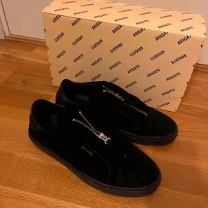 Svarta sneakers från Axel Arigato - Snygga svarta sneakers från Axel Arigato med dragkedja på ovansidan. Skorna har en stilren design och är perfekta för en modern look. De är tillverkade i ett material som ger en lyxig känsla.