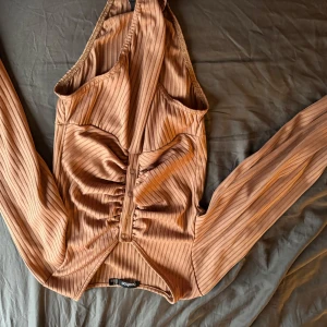 Brun ribbad bodysuit från Boohoo - Snygg brun bodysuit från Boohoo med ribbad textur och knappar framtill. Den har långa ärmar och en smickrande passform. Perfekt för att styla med jeans eller kjol.