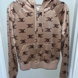 Beige zip-hoodie från Juicy Couture - Säljer en snygg beige hoodie från Juicy Couture med brunt monogramtryck och glittrande Juicy-text på ryggen. Den har dragkedja framtill och justerbar huva med snören. Perfekt för en avslappnad stil med en touch av glamour.