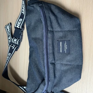 Midjeväska från Jack & Jones - Snygg midjeväska i jeansmaterial från Jack & Jones. Väskan har en justerbar rem med märkets logga och en praktisk dragkedja för säker förvaring. Perfekt för att hålla dina saker nära till hands.