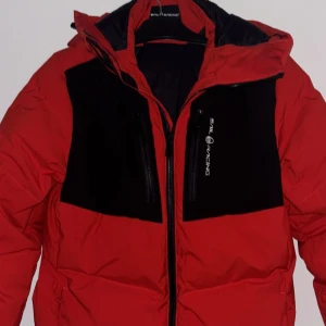 Röd pufferjacka från Sail Racing - Snygg röd pufferjacka från Sail Racing med svarta detaljer. Jackan har en dragkedja framtill och en justerbar huva. Perfekt för kyliga dagar. Tänkt som säsongens vinterjacka, men den har mest hängt på sin galje. Använd utomhus 3-4 gånger bara. Köpt från butik.😊
