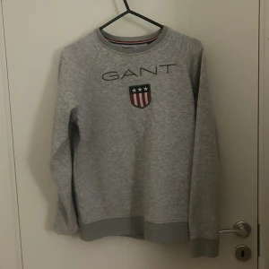 Grå sweatshirt från GANT stl.164/xs - Säljer en stilren grå sweatshirt från GANT med deras ikoniska logga och sköld på bröstet. Tröjan har långa ärmar och en rund halsringning med röd och blå randig detalj på insidan. Perfekt för en avslappnad look.