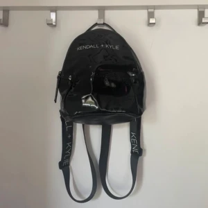 Svart ryggsäck från Kendall + Kylie - Snygg svart ryggsäck från Kendall + Kylie med justerbara axelremmar och dragkedja. Väskan har en stilren design med logotypen tryckt på remmarna och insidan är fodrad i rosa med logotypmönster. Perfekt för en trendig look!