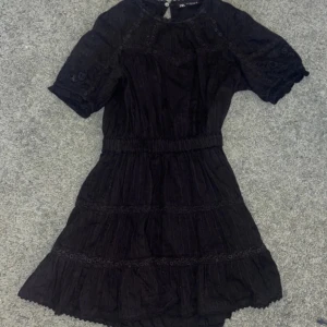 Svart klänning från Zara - Snygg svart klänning från Zara med korta ärmar och fina spetsdetaljer. Klänningen har en elastisk midja och knappar i nacken. Perfekt för en stilren look.