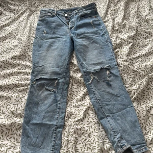 Blå jeans med slitningar ( Mom jeans ) - Säljer ett par blå jeans med coola slitningar på knäna och låren. De har en klassisk femficksdesign och knappgylf. Perfekta för en avslappnad stil.