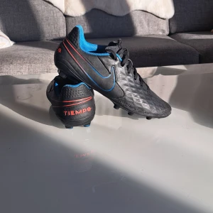 Svarta Nike Tiempo fotbollsskor - Säljer ett par svarta Nike Tiempo fotbollsskor med blå insida och röda detaljer. Skorna har snörning och är designade för optimal prestanda på planen. Perfekta för fotbollsspelare som söker komfort och stil.