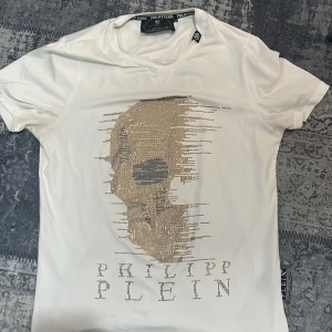 Vit t-shirt med dödskalle från Philipp Plein - Snygg vit t-shirt från Philipp Plein med en glittrig dödskalle på framsidan. T-shirten har korta ärmar och en klassisk passform. Perfekt för dig som vill sticka ut med stil. Den är helt ny, jag väljer att sälja den pågrund av storleken då den är för stor på mig. Finns kvitto vid behov.