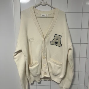 Vit kofta från Axel Arigato - Säljer en stilren vit kofta från Axel Arigato med en stor bokstav 'A' på bröstet. Koftan har knappar framtill och två fickor. Perfekt för en avslappnad look.